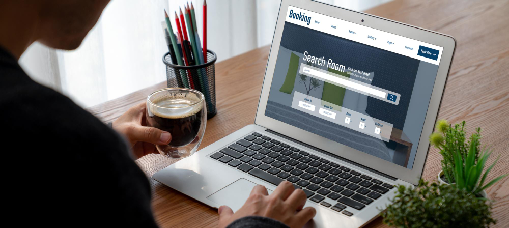 A person using a laptop displaying a booking website interface while holding a cup of coffee.